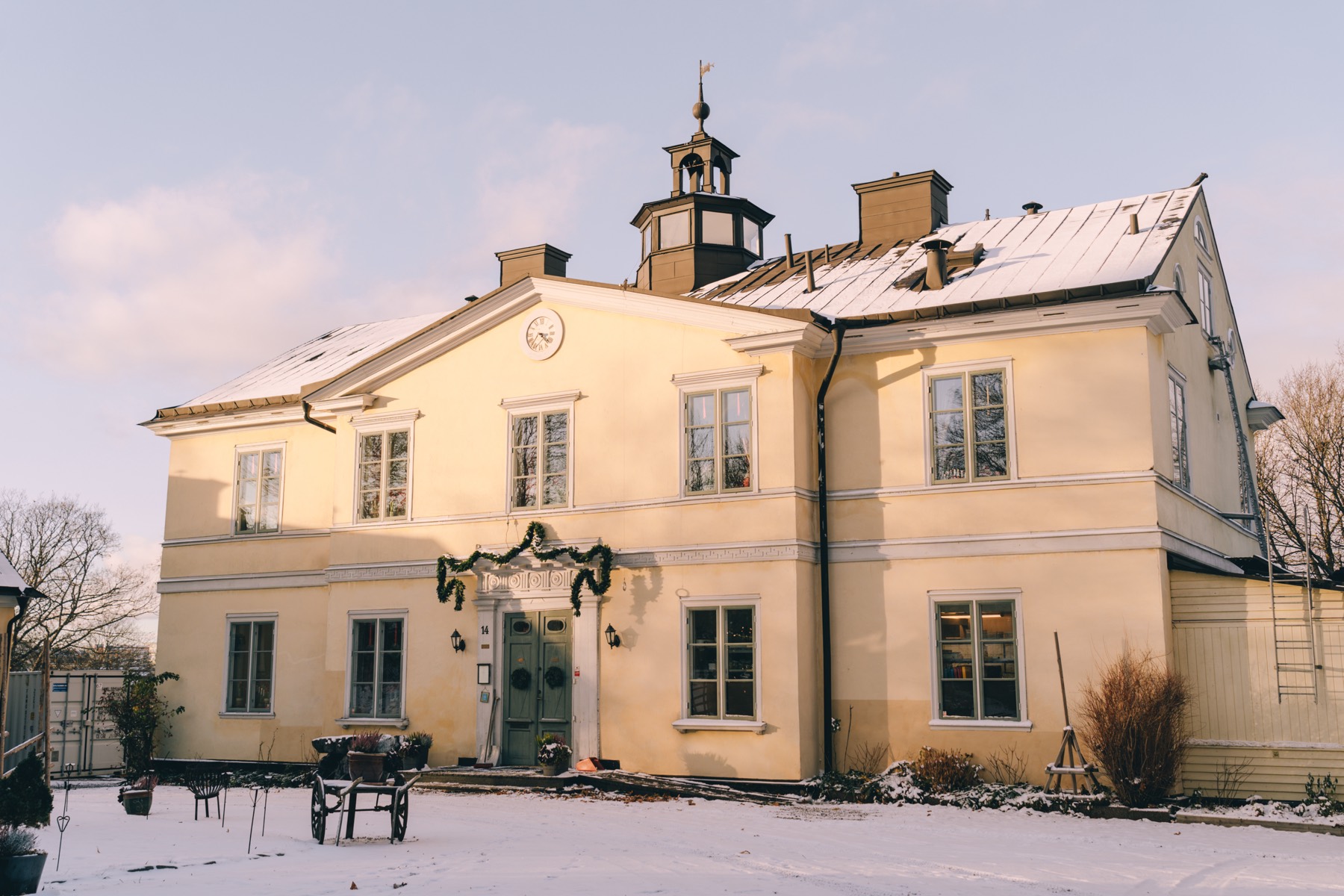 Den gula herrgårdsvillan med klocktorn i snö, girlang över porten och en gammal kärra på gårdsplanen