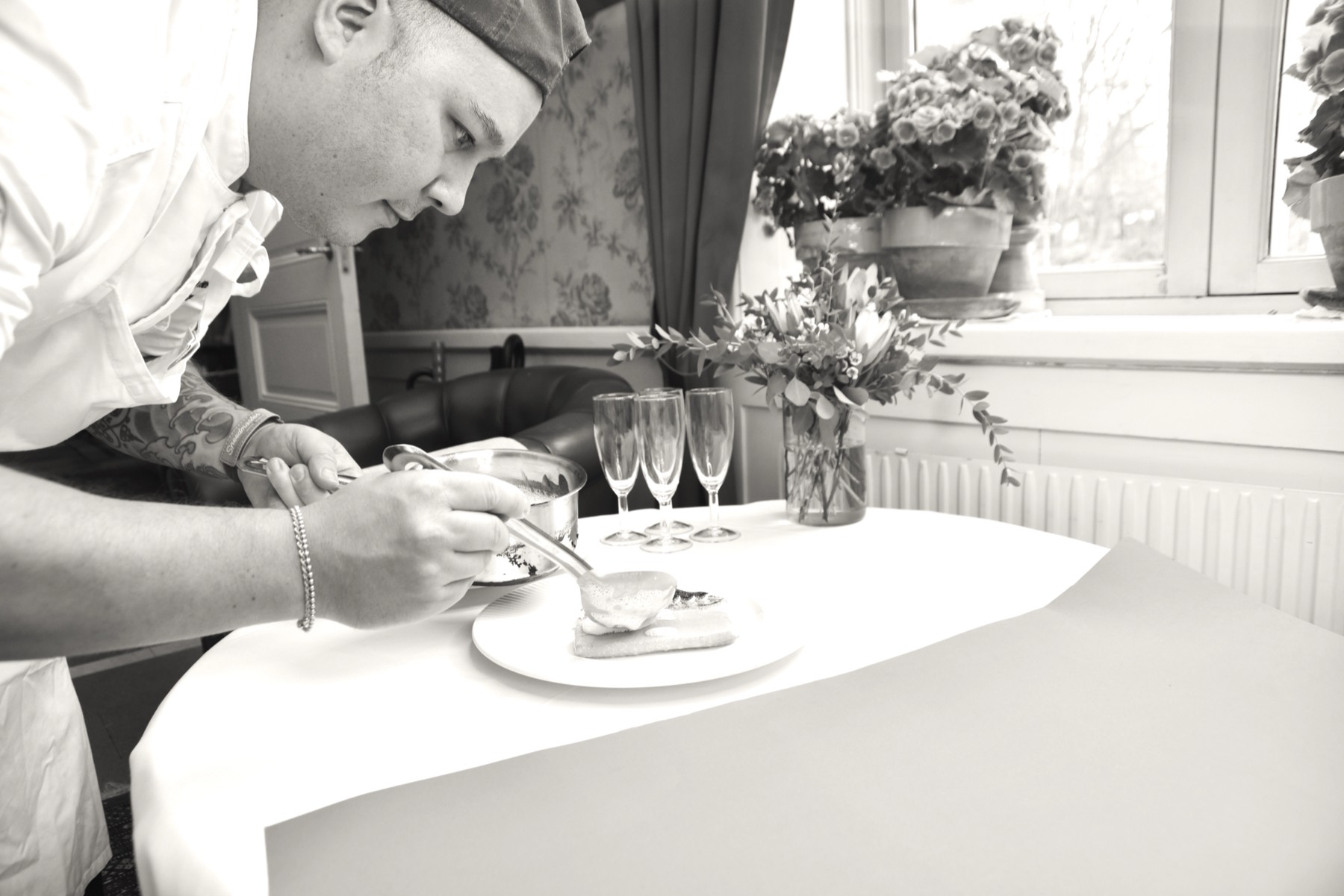 Kock i vit rock lägger upp en förrätt på tallrik vid ett vitdukat bord, med champagneglas och blommor i fönstret bakom