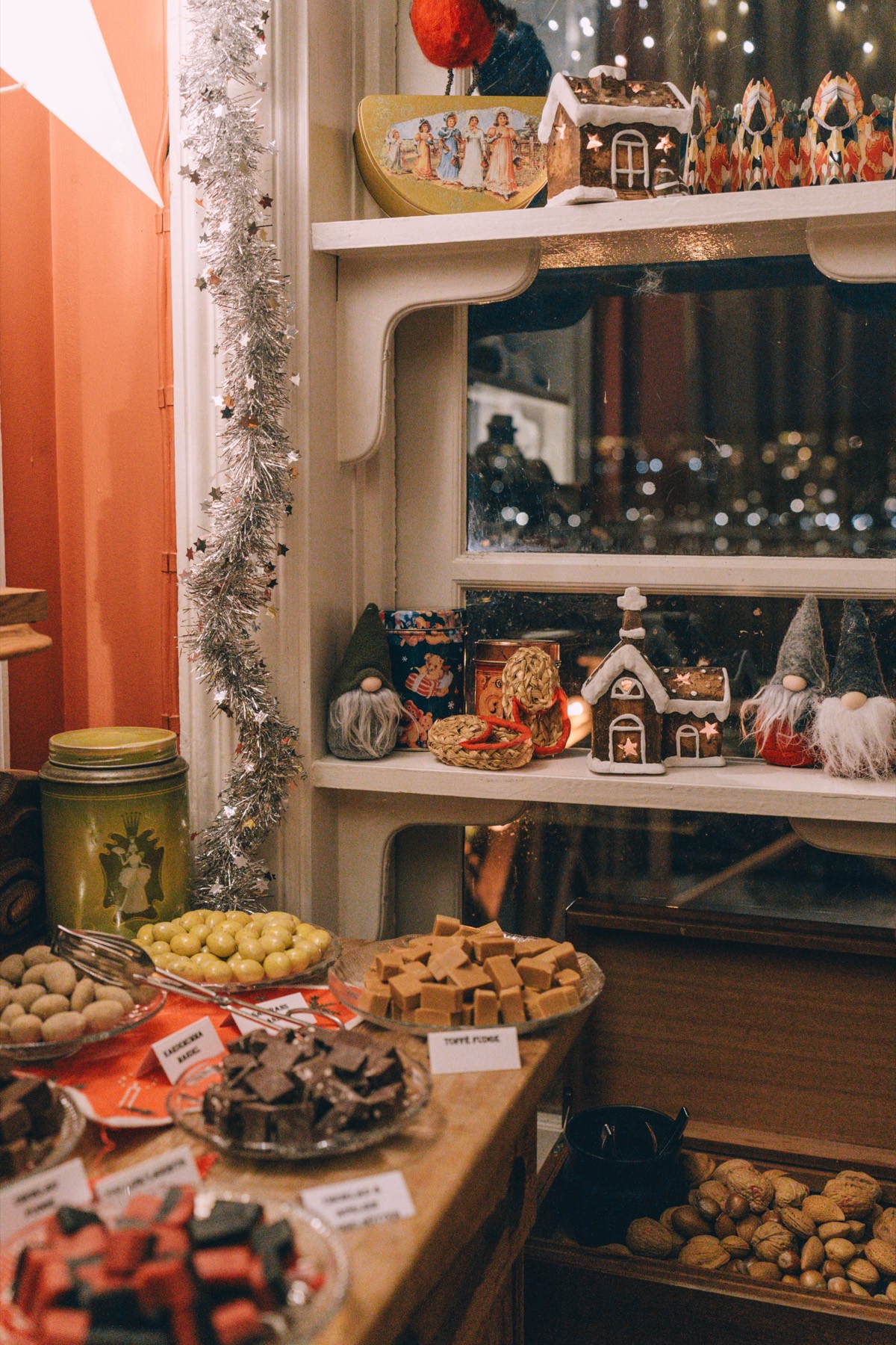 Hyllor med pepparkakshus och tomtar ovanför fat med fudge, choklad och nötter med handskrivna skyltar