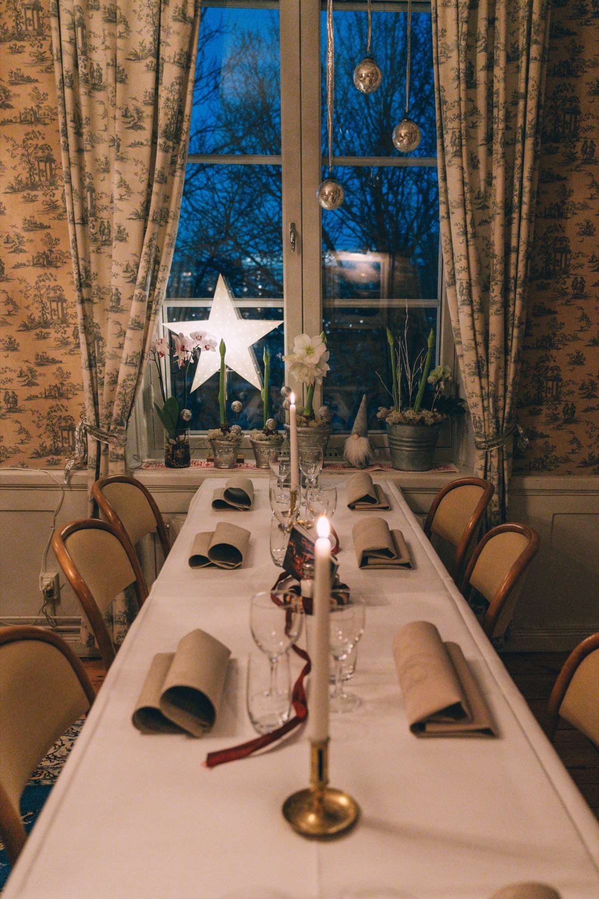 Dukat långbord med tänt ljus i mässingsstake, pappersstjärna och amaryllis i fönstret i vinterskymningen